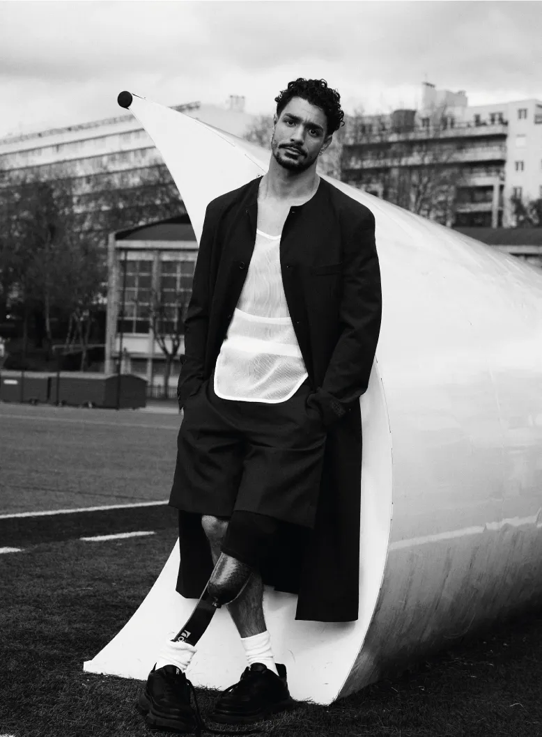 Man in stylish outfit leans against sculpture outdoors.